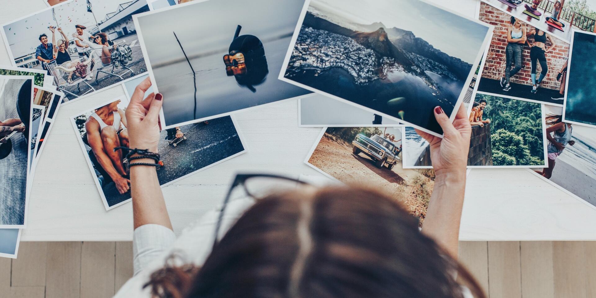 A person with long brown hair looks at a collection of colorful travel photos spread out on a white table, holding two images featuring landscapes and a cityscape.