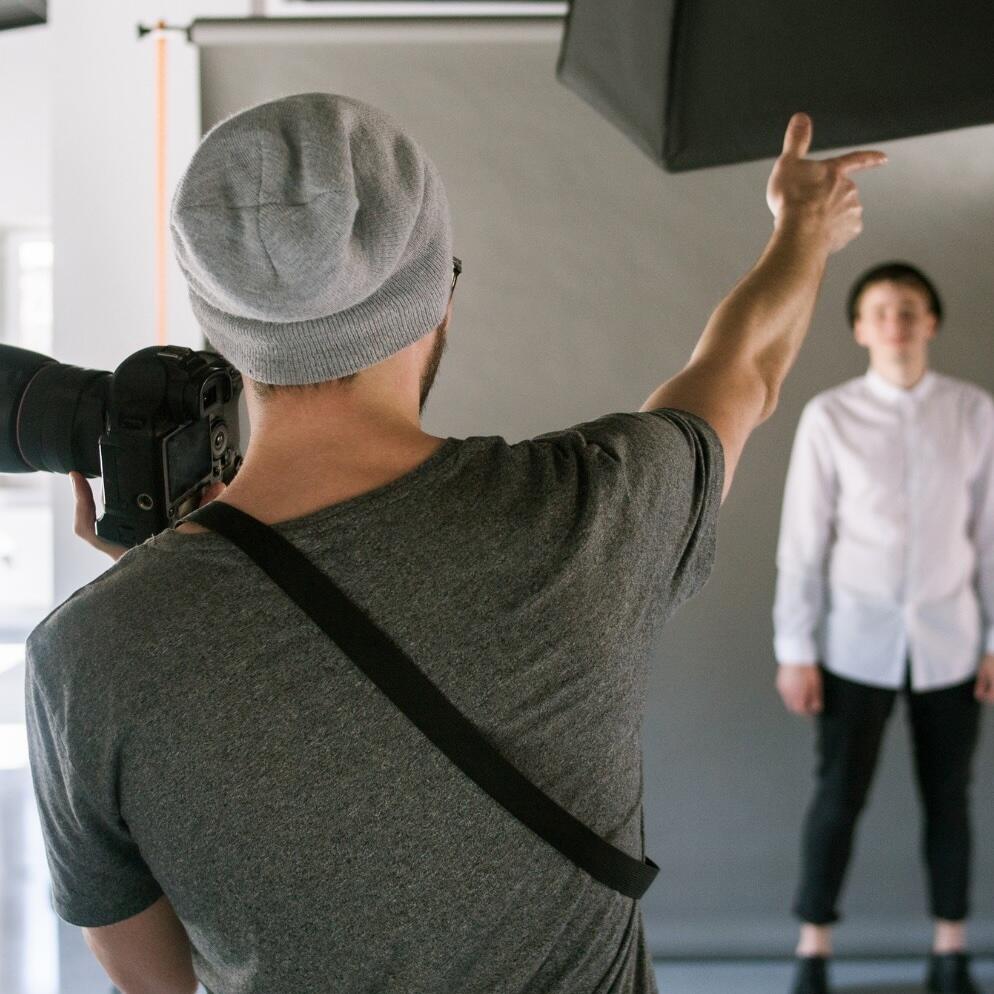 A photographer wearing a gray t-shirt and beanie points toward a person posing in a white shirt and dark pants in a studio with a gray backdrop and professional lighting.