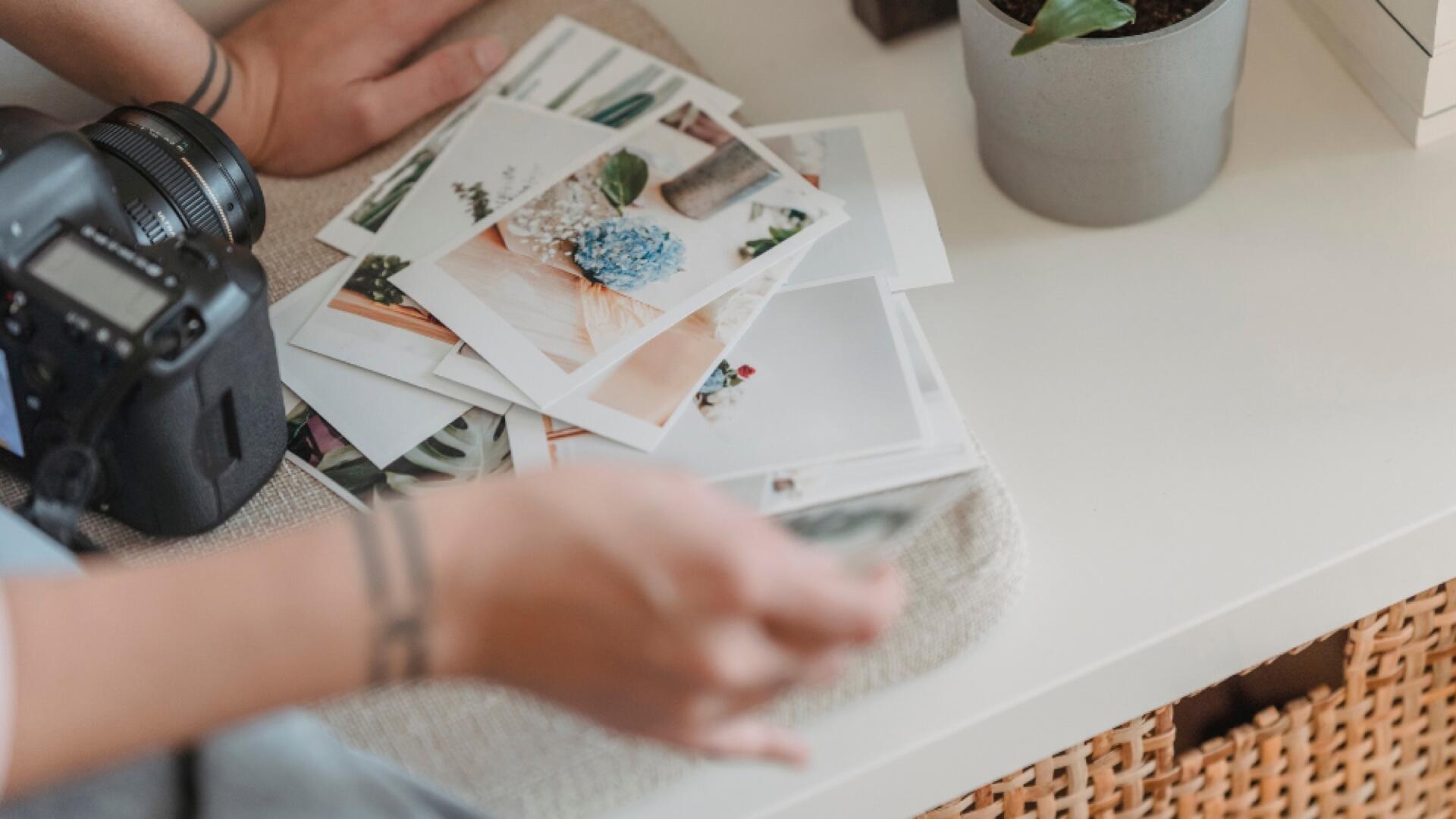 A person’s hand reaches for a stack of printed photographs on a white table next to a camera, a potted plant, and a woven drawer. The photos display plants, flowers, and nature scenes.