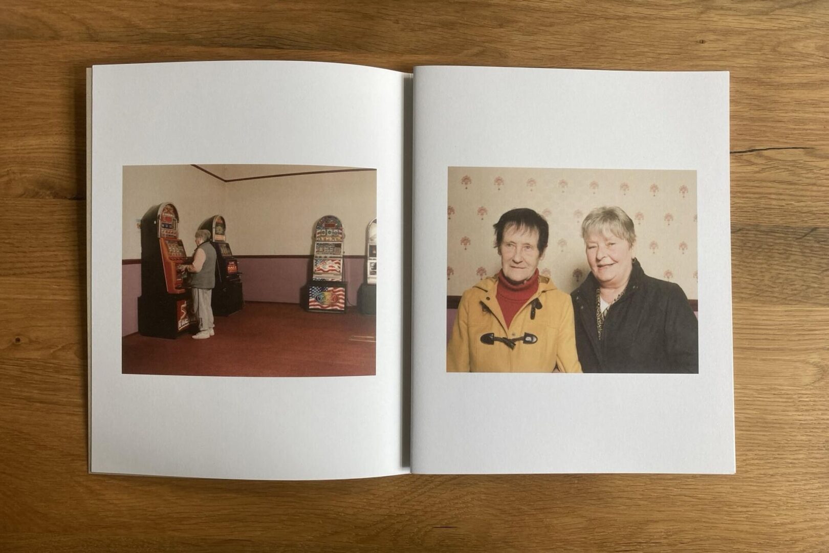 A photo book open on a wooden surface; the left page shows a person using a slot machine, and the right page features two older adults smiling and standing together in front of a patterned wall.