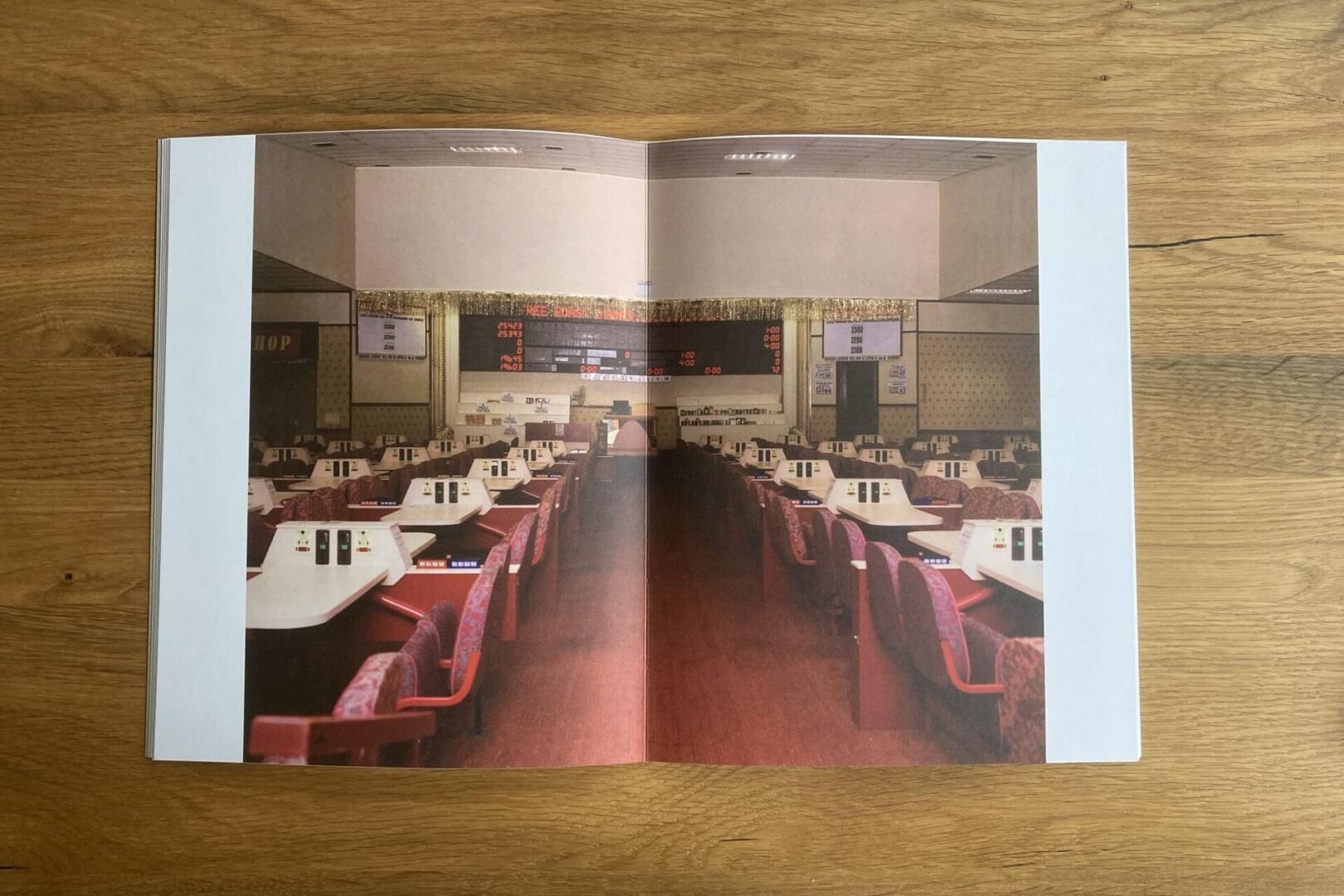 A photo of an empty bingo hall with rows of red chairs and white tables, each with bingo cards and markers. Digital boards display numbers at the front, and the room has wood-paneled flooring and beige walls.