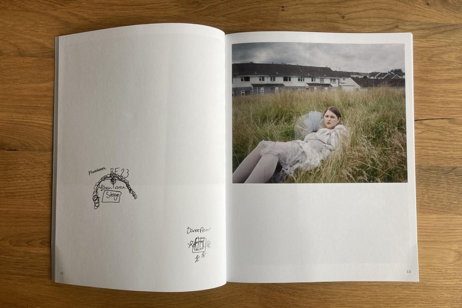 An open magazine on a wooden table shows a photo of a young woman lying in tall grass with houses in the background on the right page, and simple black ink sketches on the mostly blank left page.