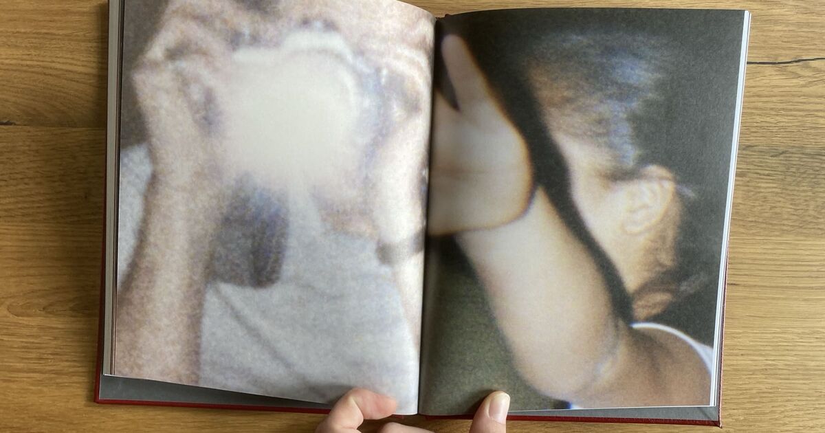 A blurry photo in an open book shows one person taking a picture with a flash on the left page, while another person on the right page raises their hand, partially blocking their face. The book is held open on a wooden surface.