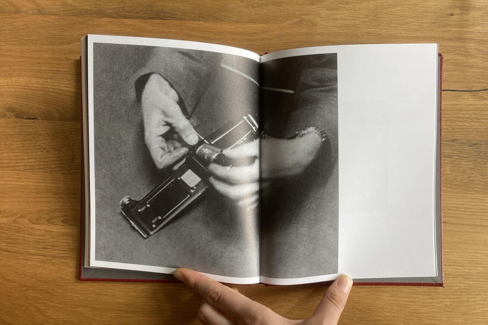 An open book rests on a wooden surface. The left page shows a black-and-white photo of hands holding a camera or mechanical device. A hand holds the book at the bottom center. The right page is mostly blank.