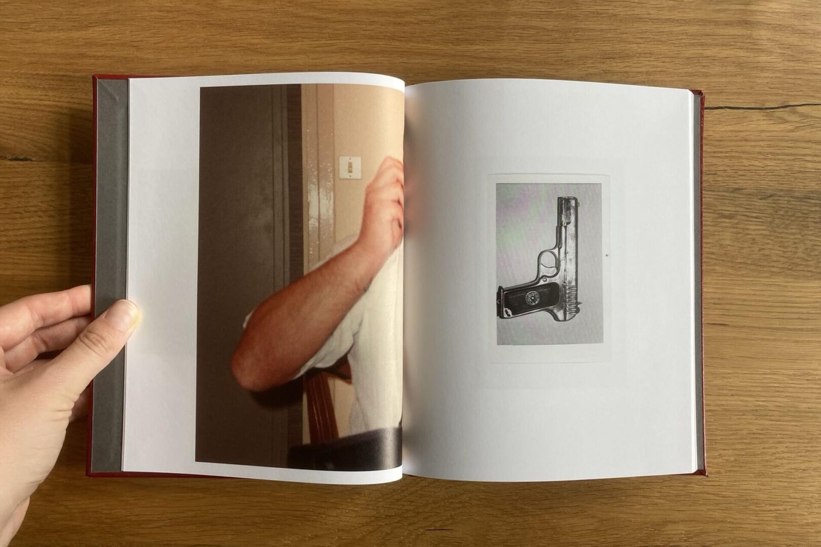 An open book shows a cropped photo of a person’s arm and torso on the left page and a black-and-white illustration of a handgun on the right page. A hand holds the book open on a wooden surface.