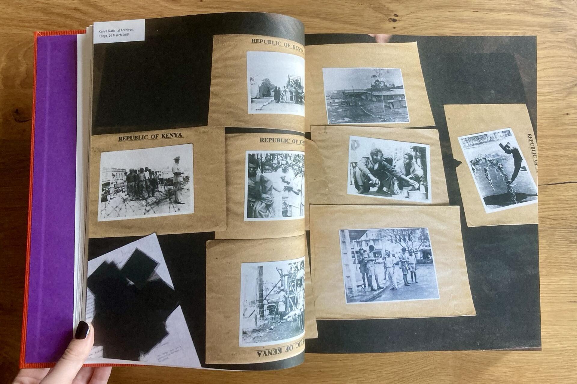 A person holds open a scrapbook displaying multiple vintage black-and-white photos, some labeled Republic of Kenya, arranged on black pages with tan photo corners. The scrapbook rests on a wooden surface.