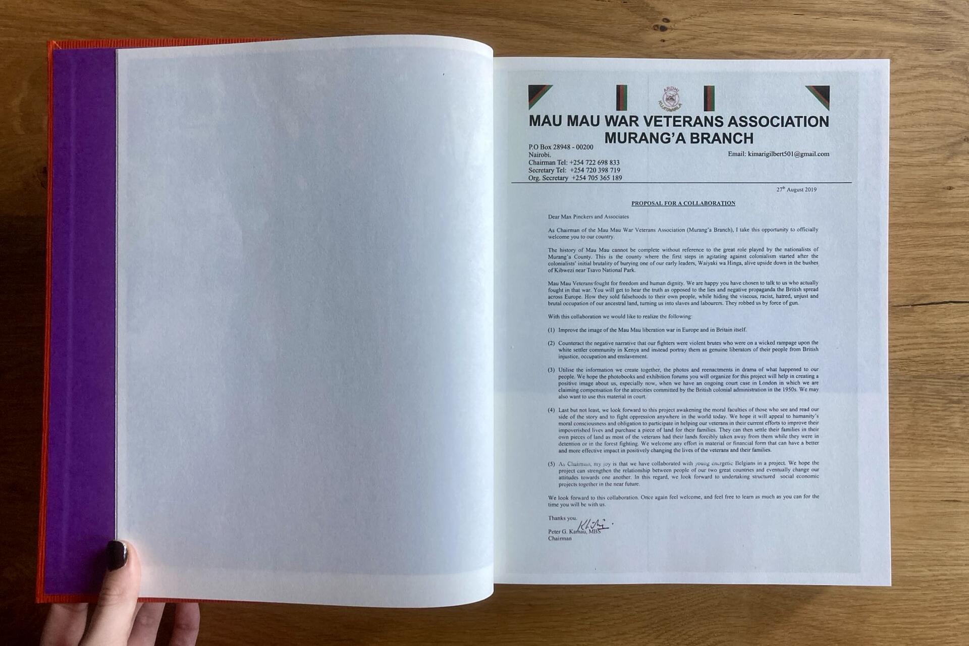 A person holds open a book with a blank left page and a right page showing a formal letter from the MAU WAR VETERANS ASSOCIATION MURANGA BRANCH on headed paper, placed on a wooden surface.