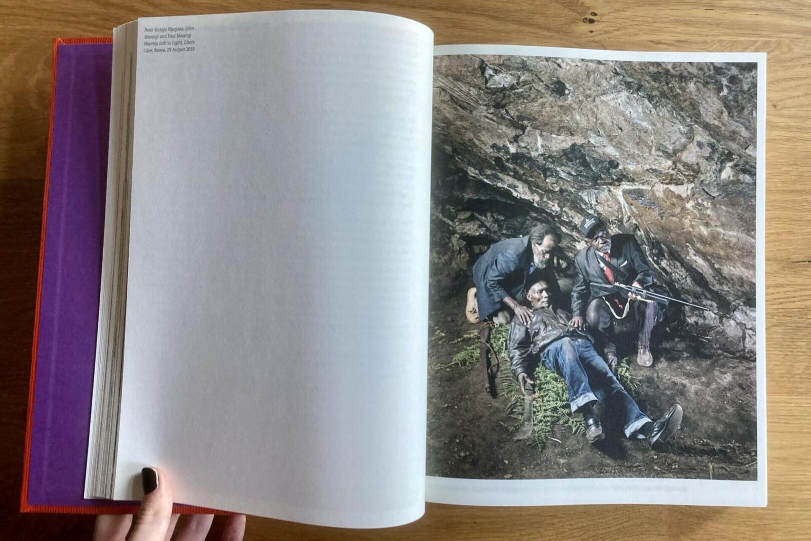 A person holds open a book. The left page is mostly blank, while the right page shows a photograph of three people sitting and lying on rocky ground, near a rock wall, with some green plants nearby.