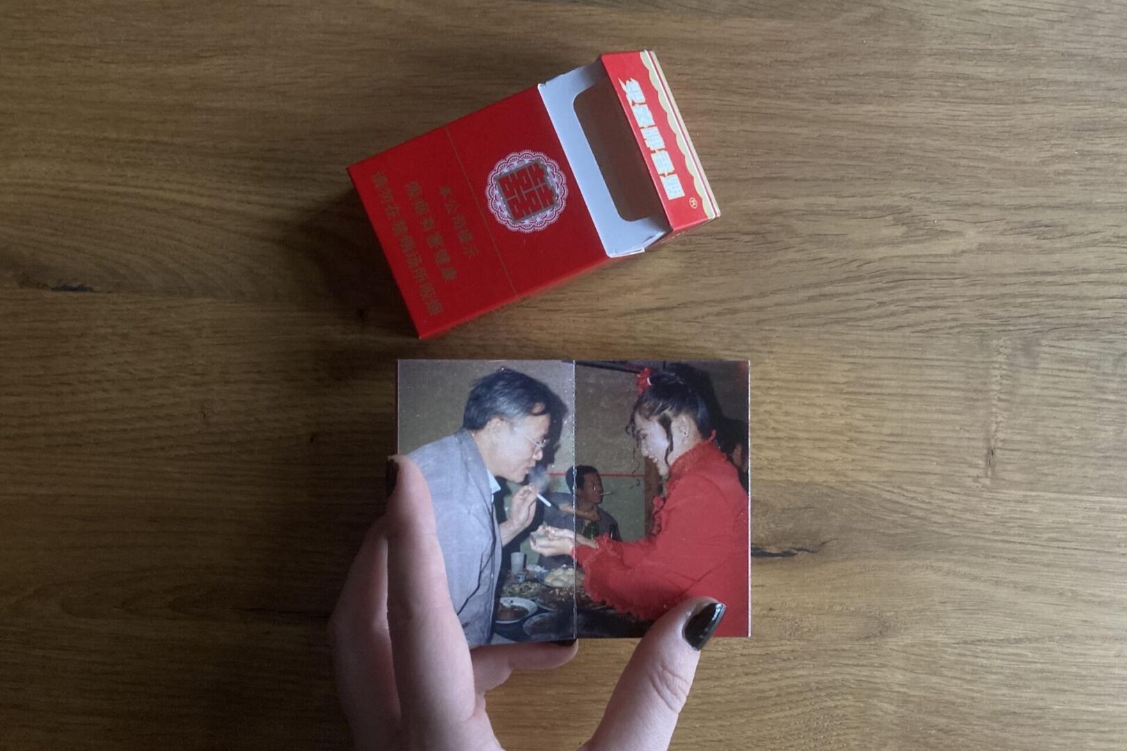 A hand with dark nail polish holds an old photo of a man and woman eating together. Above it, there is a red Hot Pot Matchbox on a wooden table.