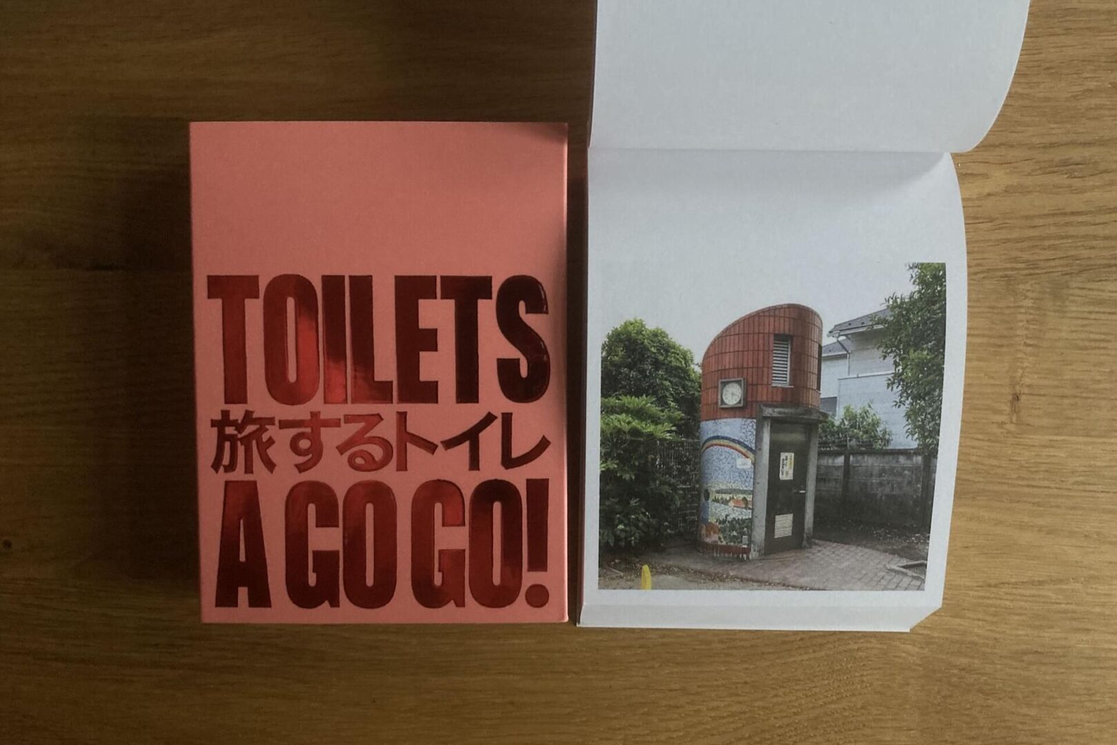A book titled TOILETS 旅するトイレ A GO GO! lies open on a wooden surface, showing a photo of a small, modern public toilet building surrounded by greenery.
