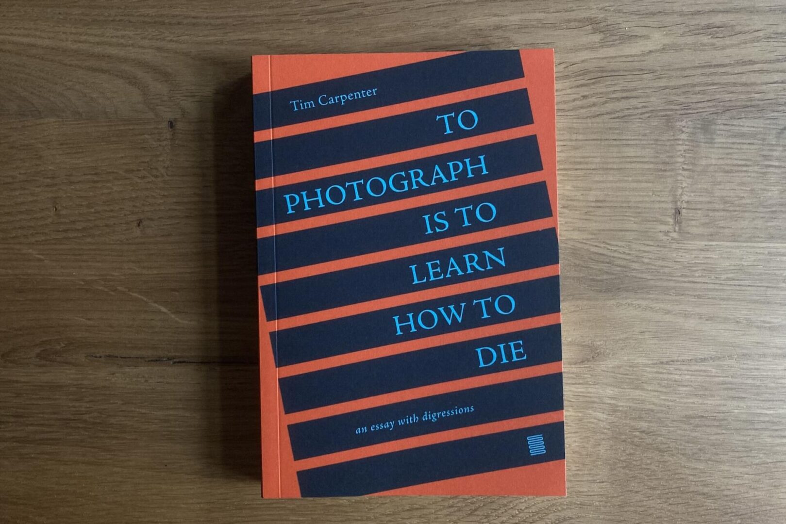 A book titled To Photograph Is to Learn How to Die by Tim Carpenter lies on a wooden surface. The cover is orange with black and blue diagonal stripes displaying the title and subtitle, an essay with digressions.