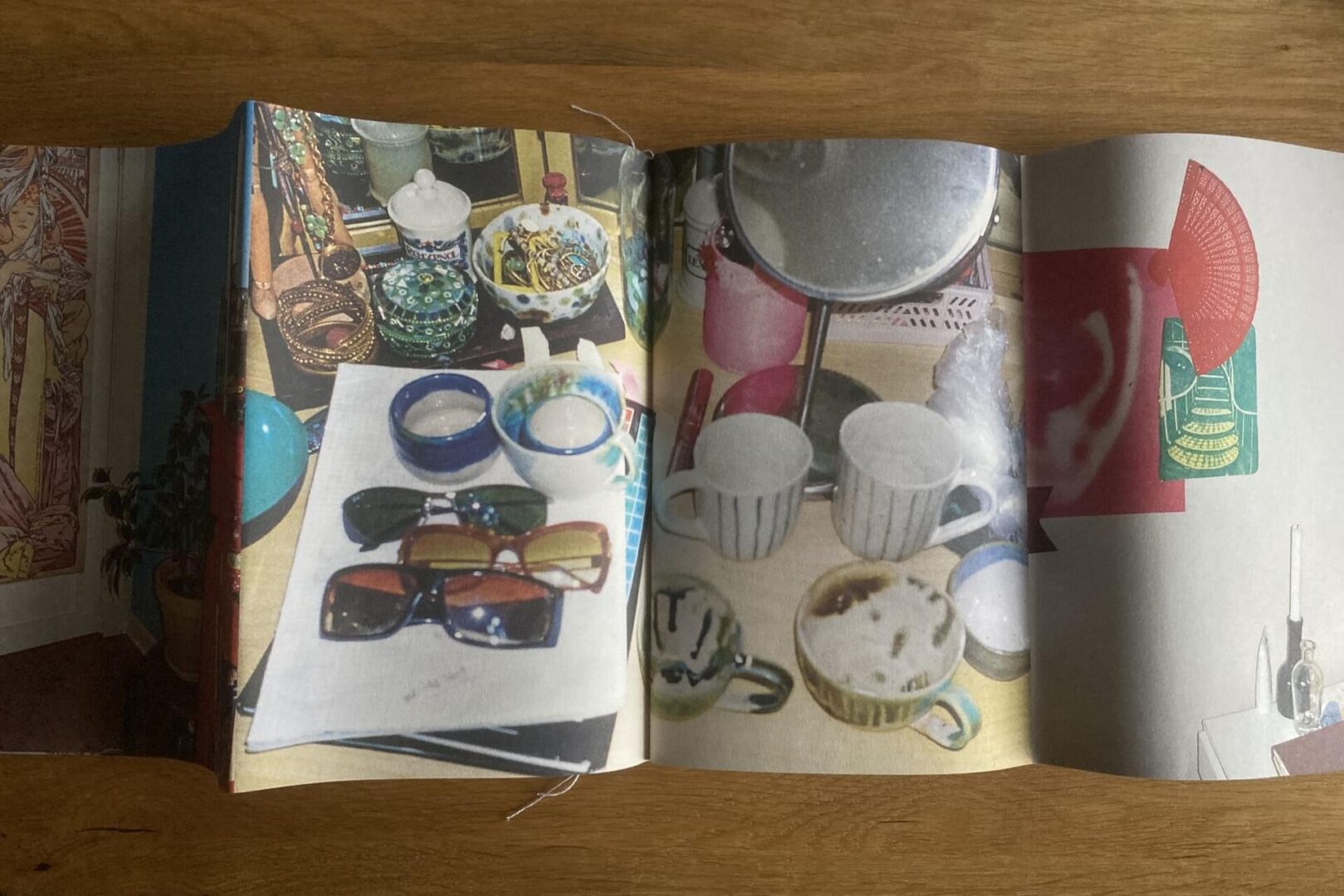 A folded booklet is open on a wooden surface, displaying colorful photos of assorted ceramic cups, bowls, sunglasses, and decorative objects on tabletops. Red, white, and green graphic elements appear on the right page.