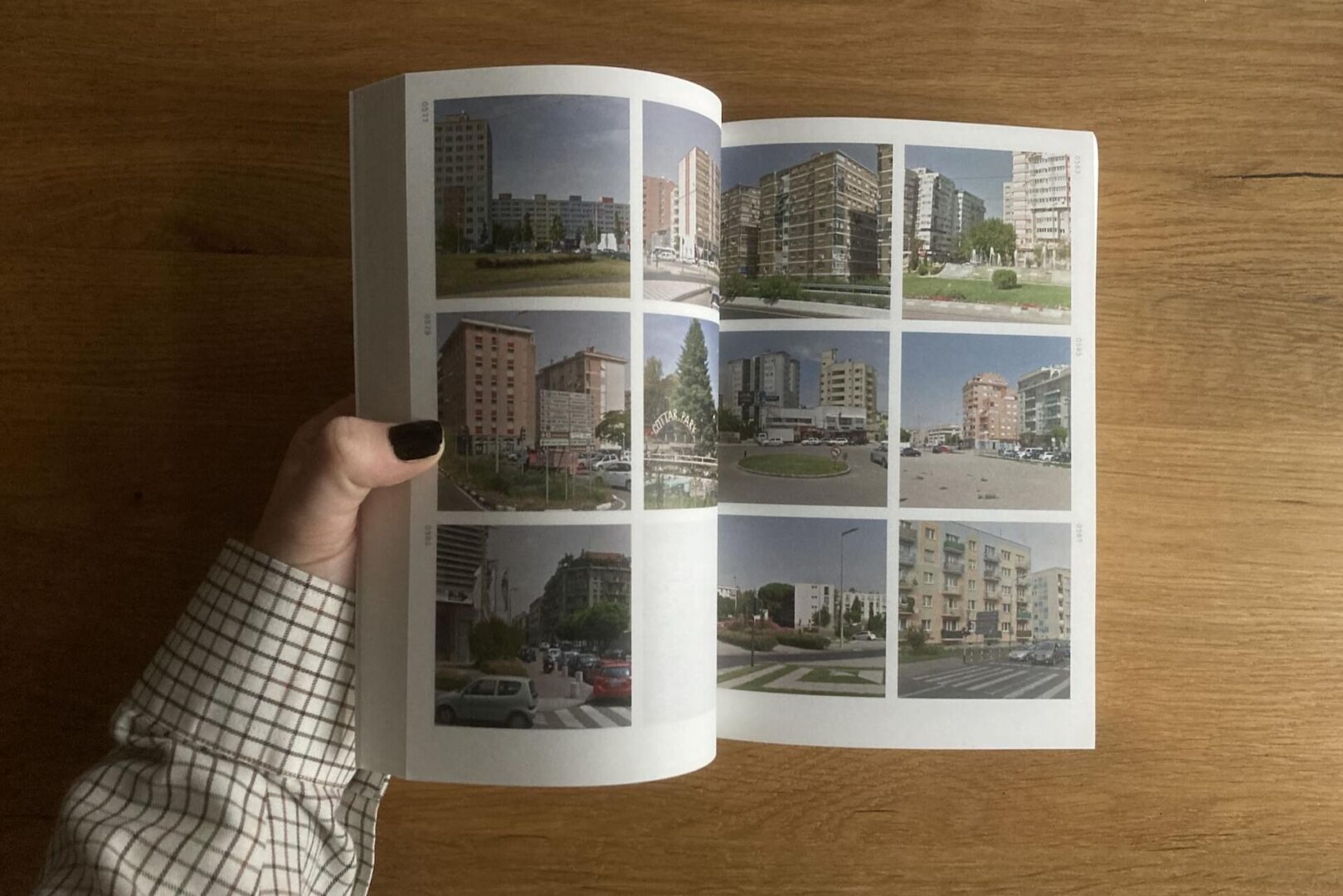 A person with dark nail polish and a checkered shirt holds open a book displaying a grid of nine photos of urban apartment buildings on a wooden table.