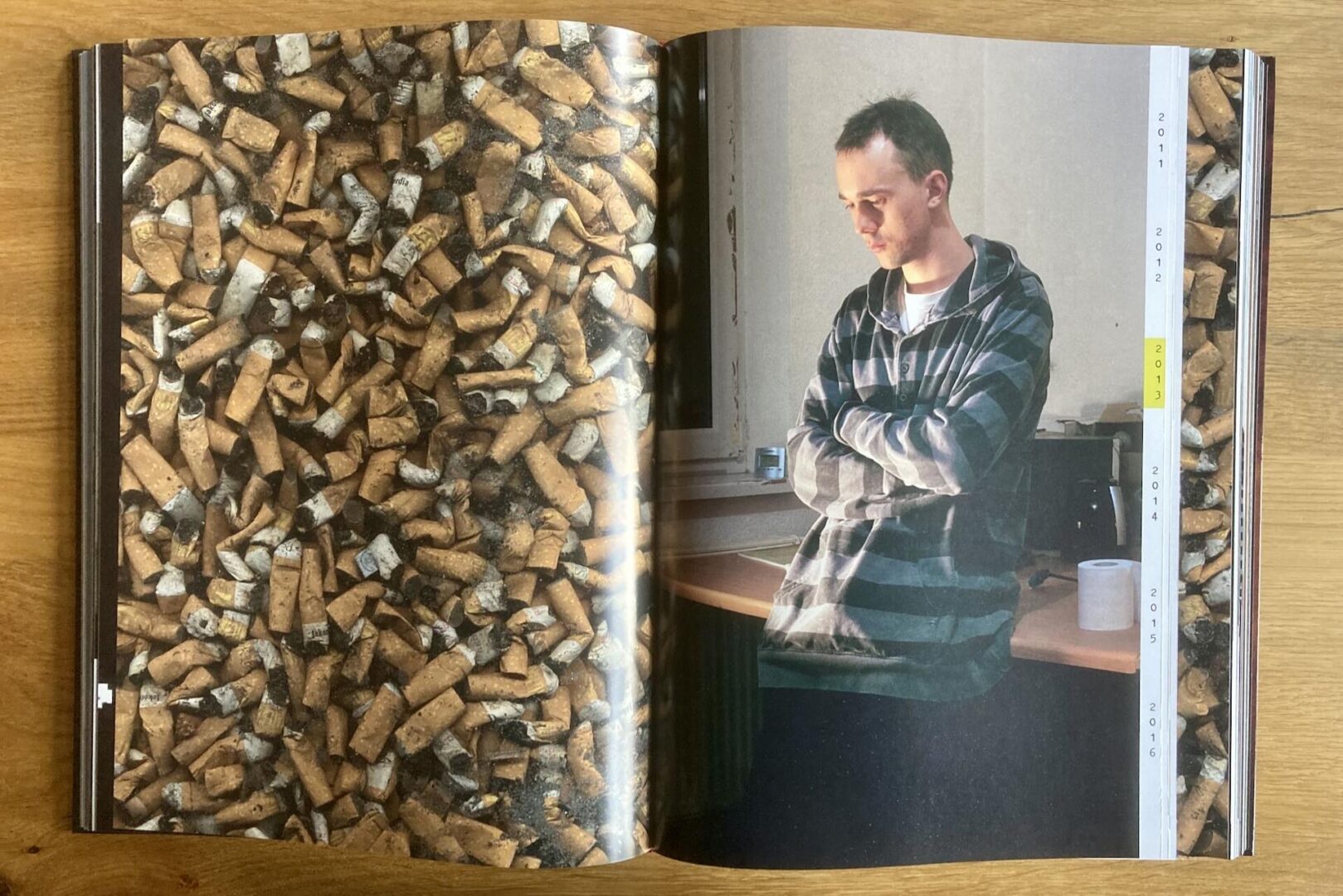 A book spread shows a left page filled with a close-up of many used cigarette butts. The right page features a young man in a striped hoodie, standing indoors with arms crossed, looking down pensively.