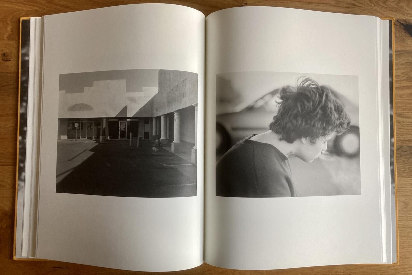 An open photo book on a wooden surface displays two black-and-white images: one of an empty building exterior in sunlight, and another of a person in profile exhaling visible breath or smoke.