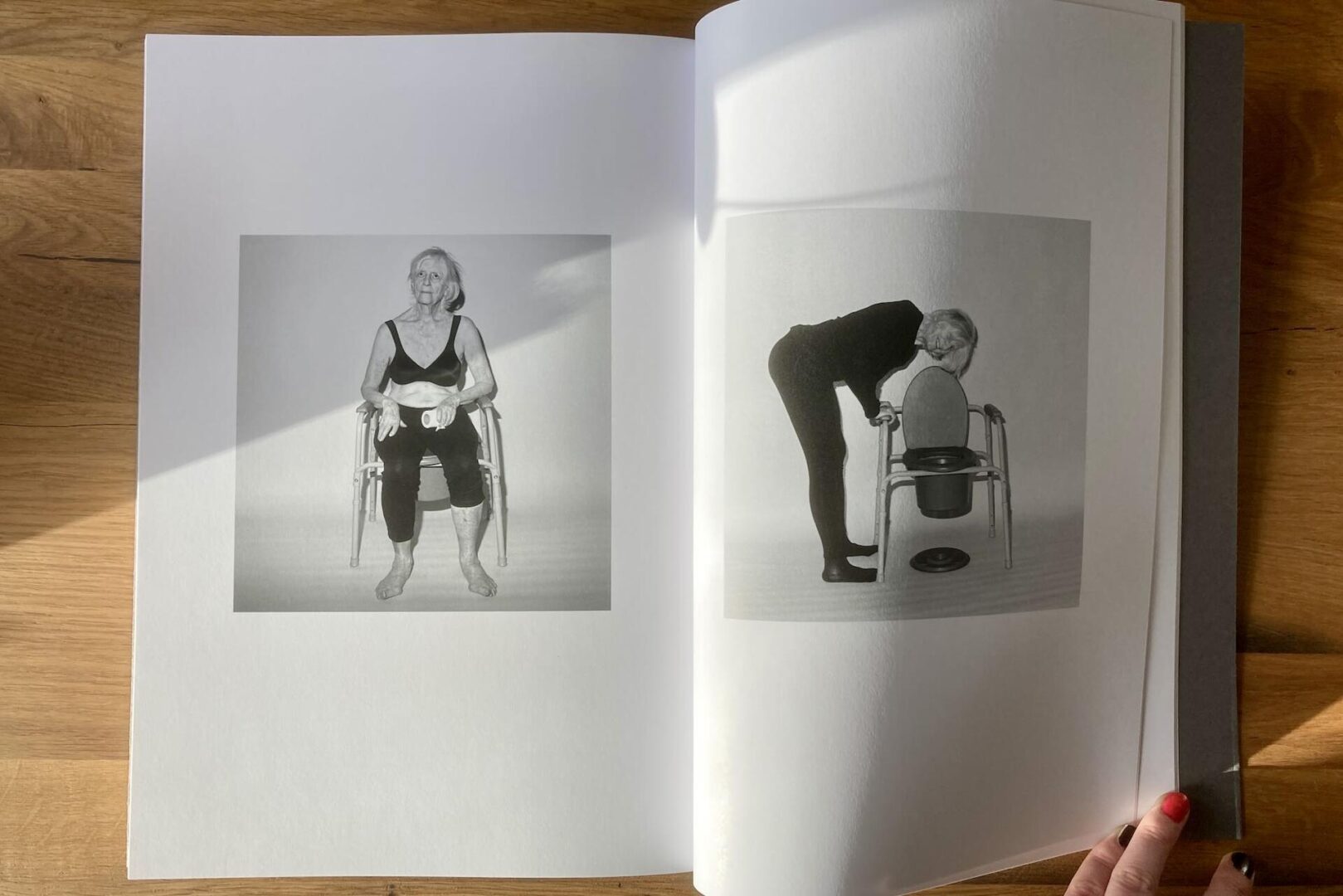 Two black-and-white photos of an older woman: on the left, she sits on a chair holding her knees; on the right, she bends forward to open the lid of a commode chair. A hand holds the book open on a wooden surface.