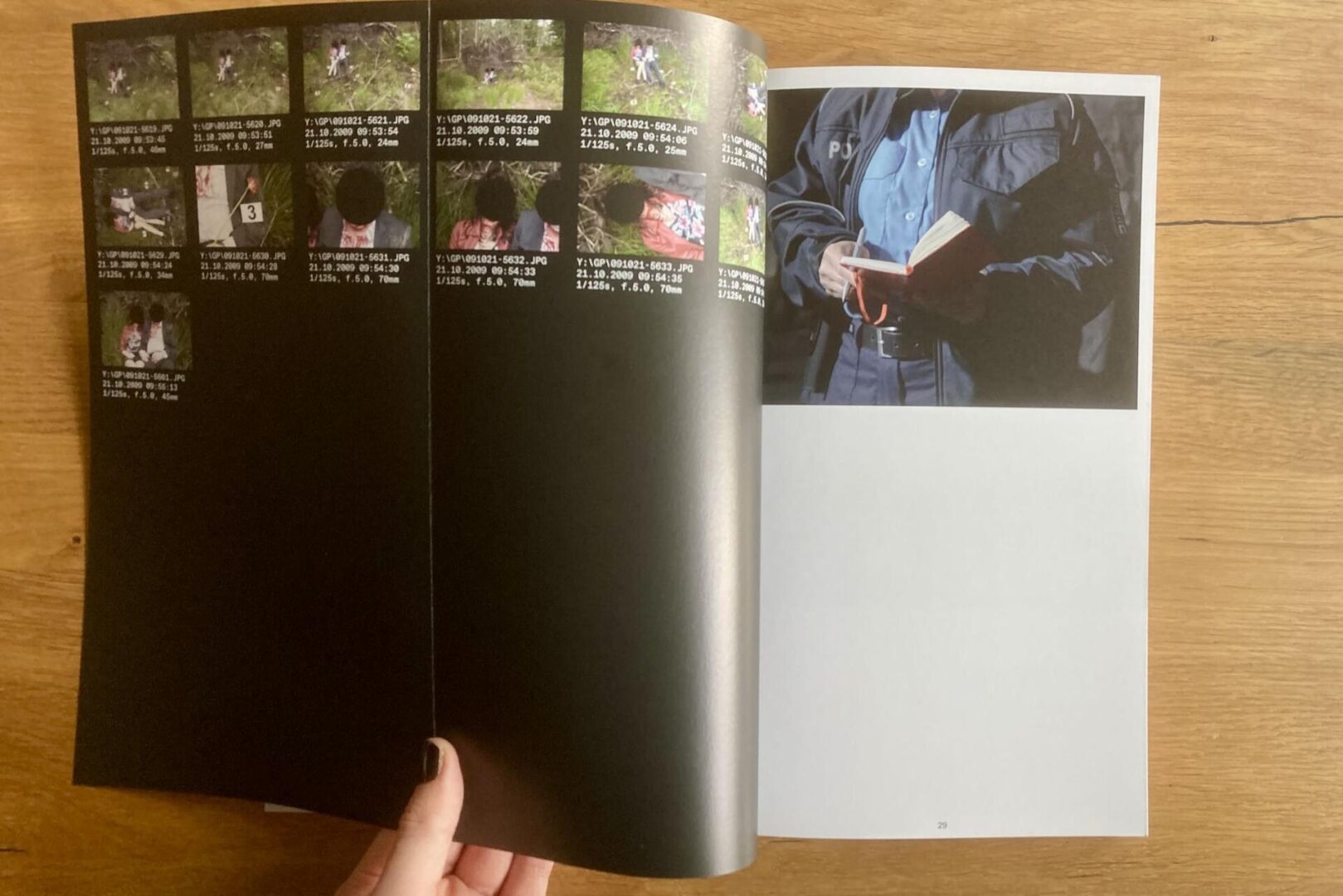 A hand holds open a magazine. The left page shows multiple small photos with captions, while the right page features a large image of a person in a dark jacket reading a book, against a plain background on a wooden table.