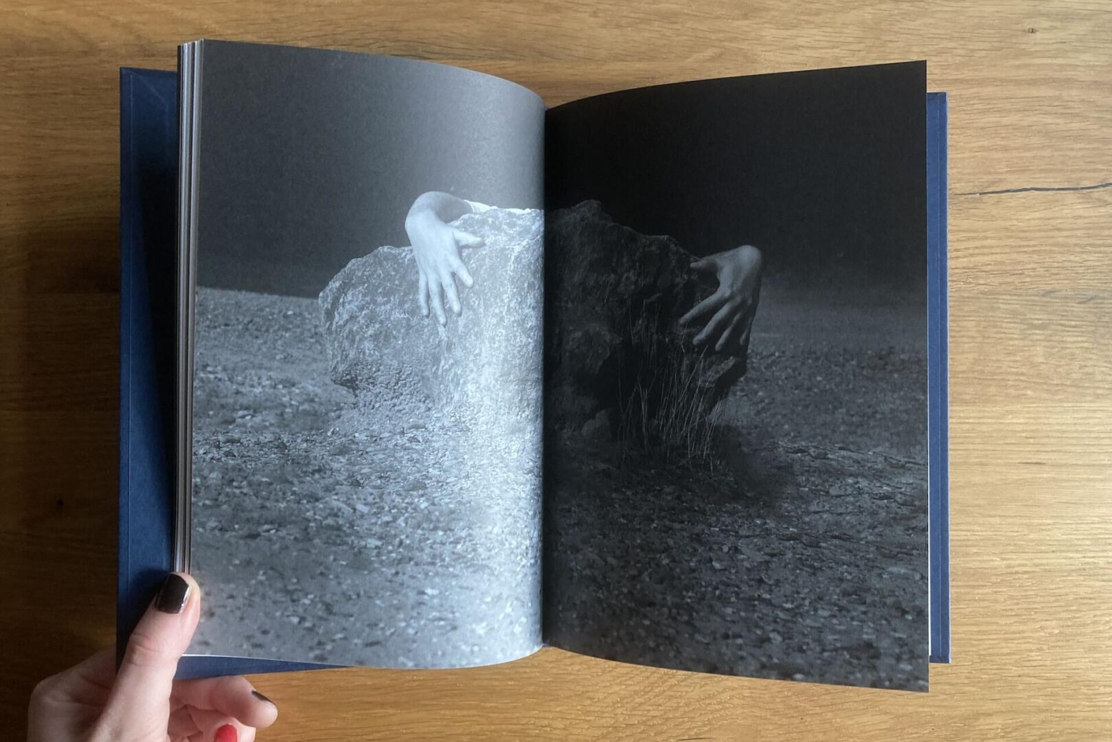A hand holds open a book. The pages show a black-and-white photograph of a large rock on a barren ground, with two human arms reaching out from behind it.