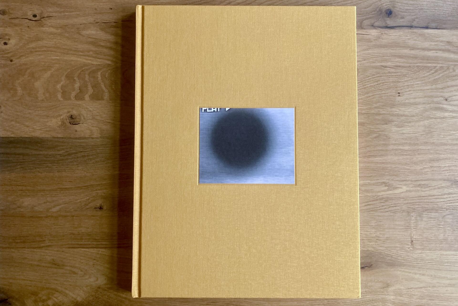 A yellow hardcover book with a small, centered cover image showing a dark, blurry circle on a gray background, placed on a wooden surface.