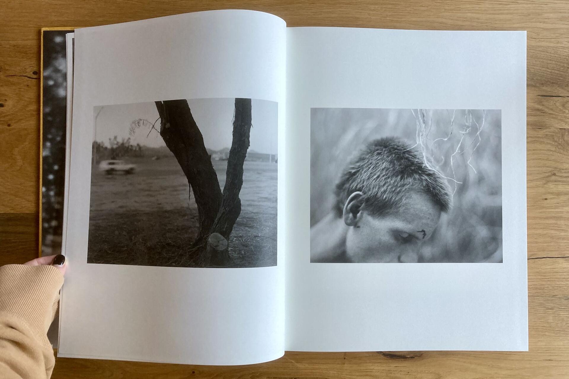 An open photo book displays two black and white images: the left page shows two trees with blurred scenery in the background; the right page features a close-up of a person looking downward, partially obscured by shadows.