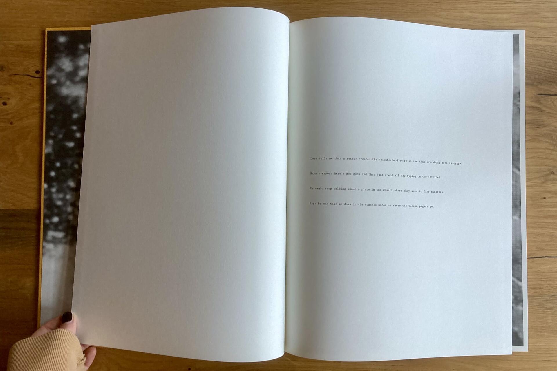 A person holds open a large book displaying a mostly blank white page with a short block of centered text, photographed on a wooden surface with blurred black-and-white images visible at the page edges.