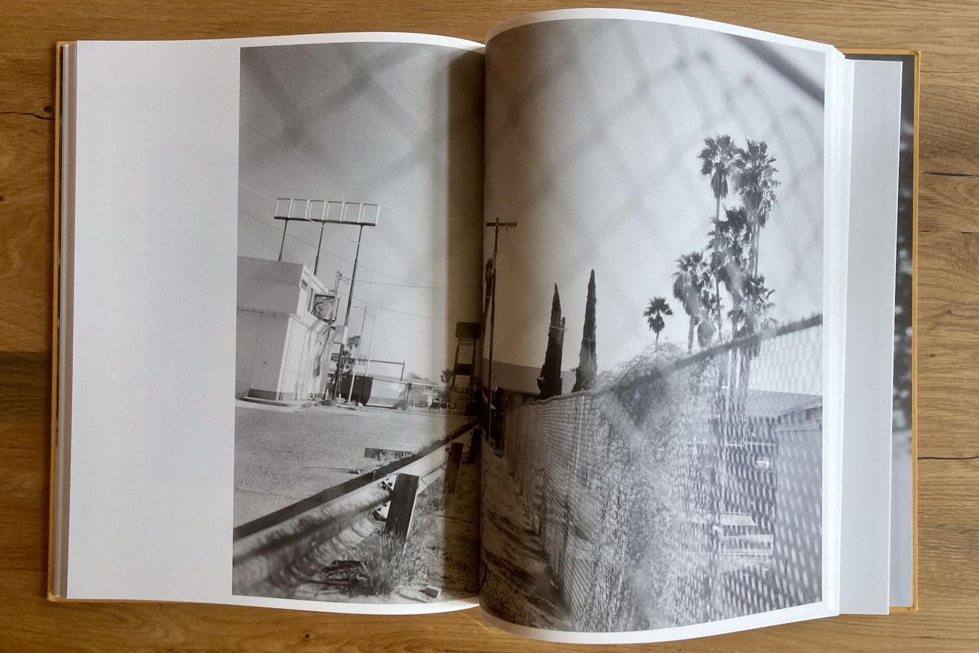 An open book displays black and white photos of a deserted street and palm trees behind a chain-link fence, with the fence pattern visible over the right page. The book rests on a wooden surface.
