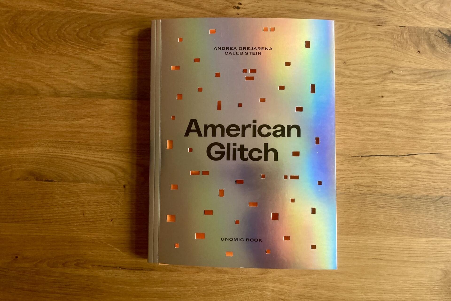 A book titled American Glitch with a shiny, holographic cover featuring square cutouts and black text, placed on a wooden surface. Authors listed are Andrea Orejarena and Caleb Stein.