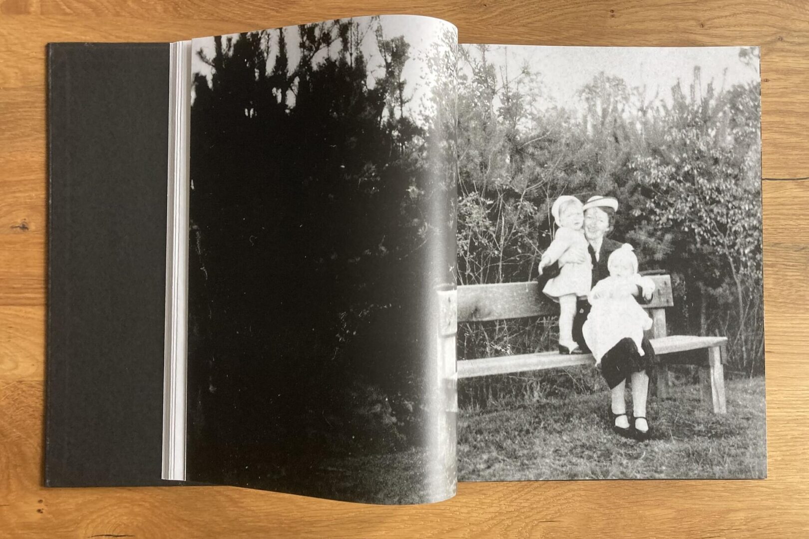 An open photo book lies on a wooden surface, showing a black-and-white image of a person sitting on a bench outdoors with two young children on their lap, surrounded by bushes.