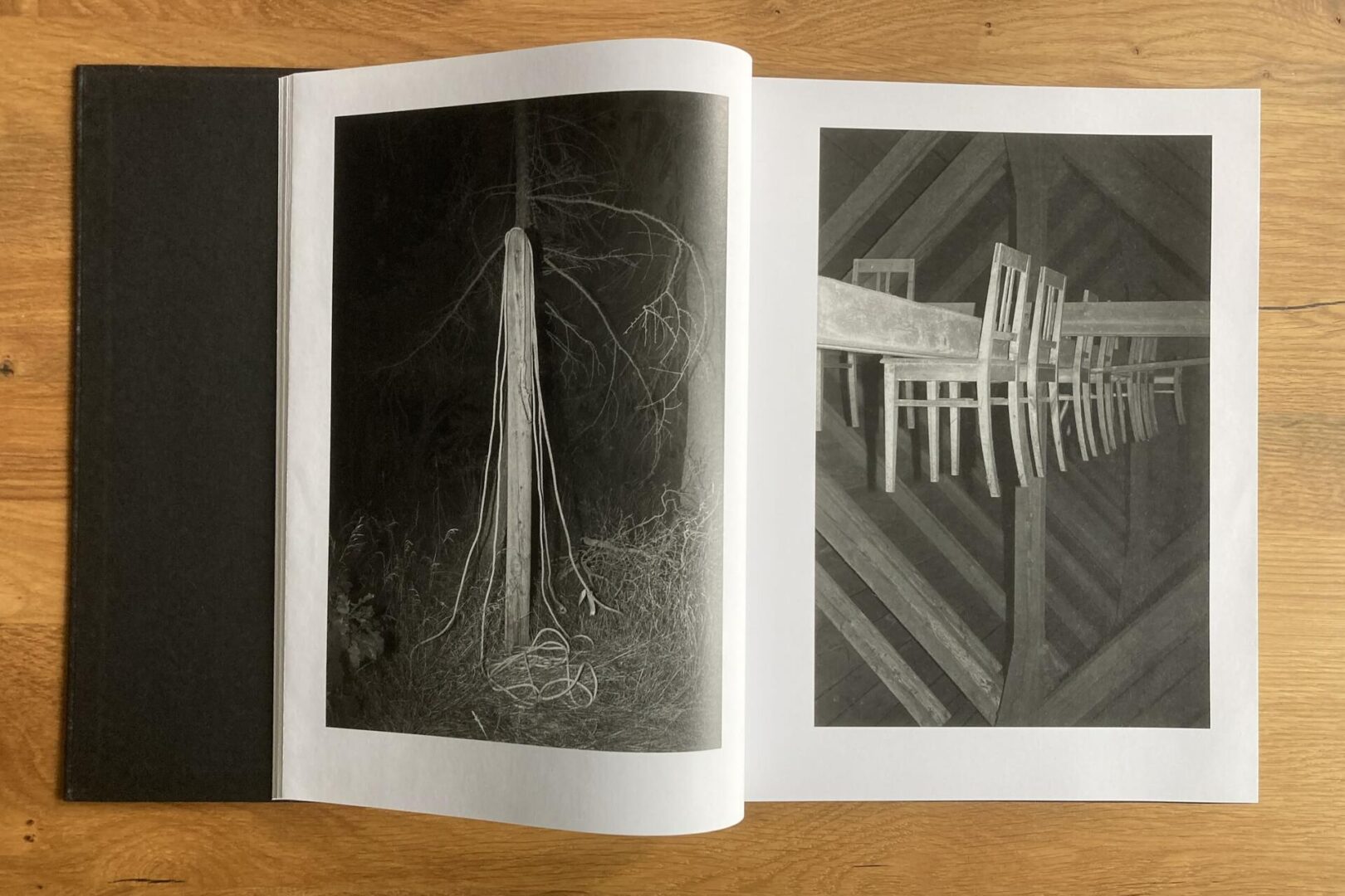 An open book on a wooden surface shows two black and white photos: one of a tree trunk with hanging ropes, and one of chairs stacked on beams in an attic-like space.