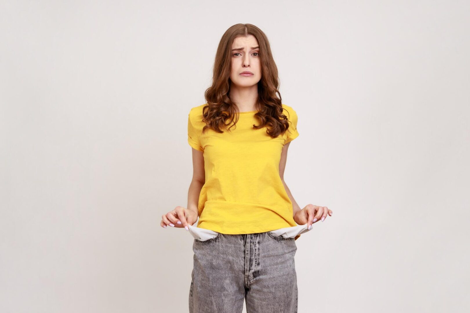 A young woman with long brown hair wears a yellow t-shirt and grey jeans, standing against a plain background. She looks worried and pulls out her empty pockets, suggesting she has no money.