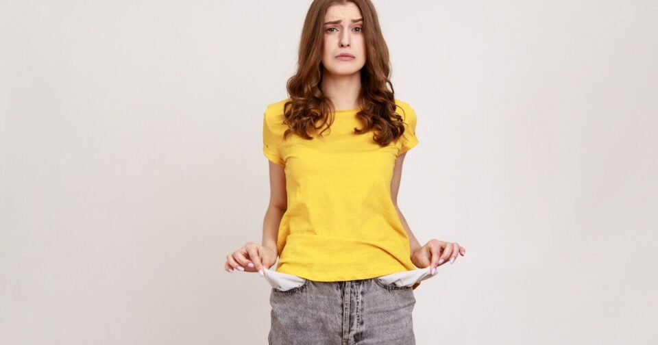 A young woman in a yellow t-shirt and gray jeans stands against a plain background, holding out her empty pants pockets with a worried expression.
