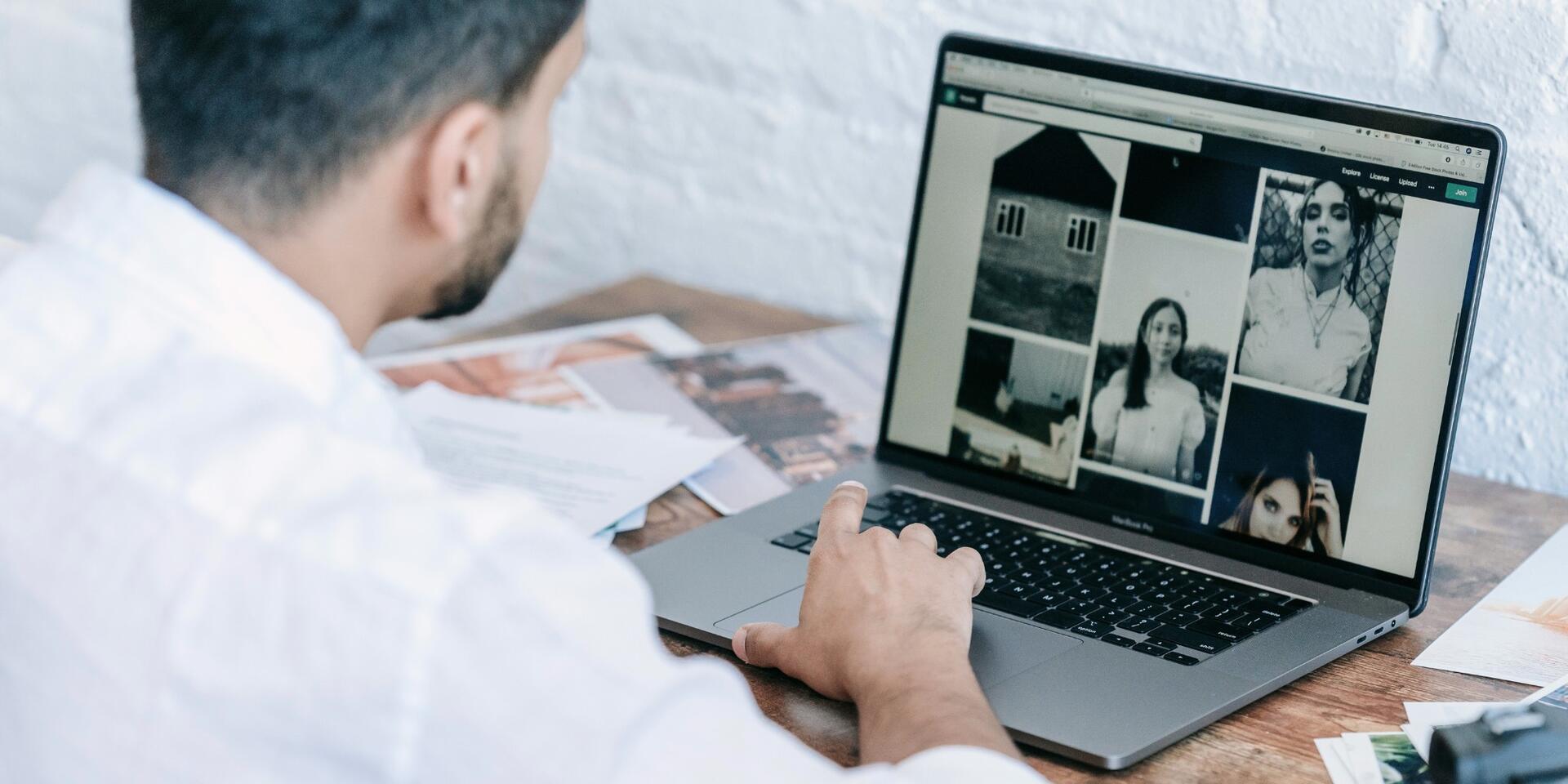 A person in a white shirt sits at a wooden desk, looking at a laptop displaying a collage of black-and-white photos, with papers scattered around.