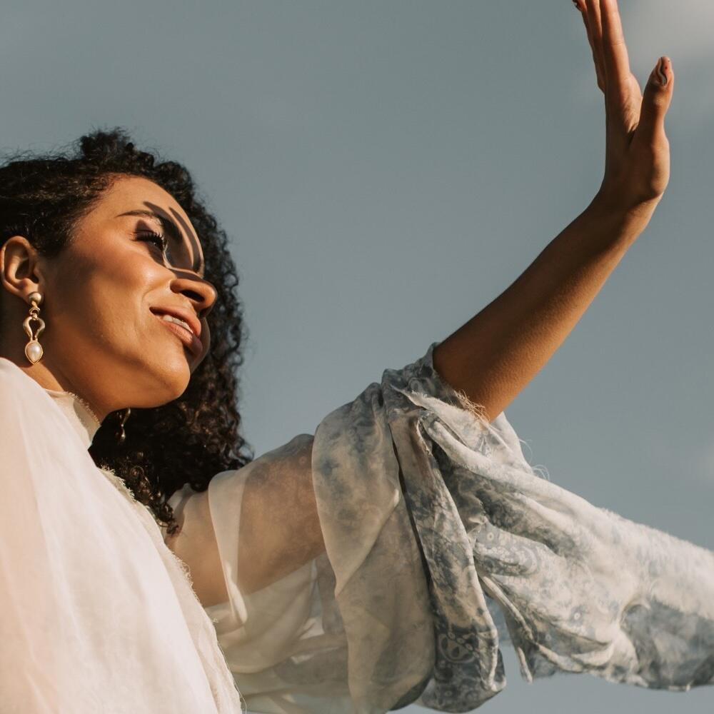 A woman with curly hair wearing a sheer, patterned sleeve and pearl earrings looks up, smiling, as she raises her arm toward the sky in sunlight.