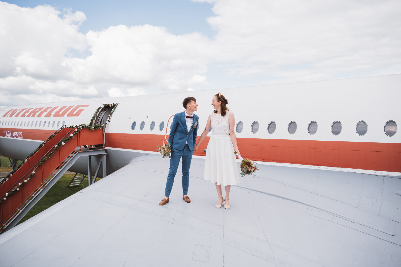 Ein Hochzeitspaar steht Hand in Hand auf der Tragfläche eines Flugzeugs und lächelt sich an. Im Hintergrund sind die Flugzeugkabine mit rot-weißer Lackierung, eine geschmückte Treppe und ein bewölkter Himmel zu sehen.