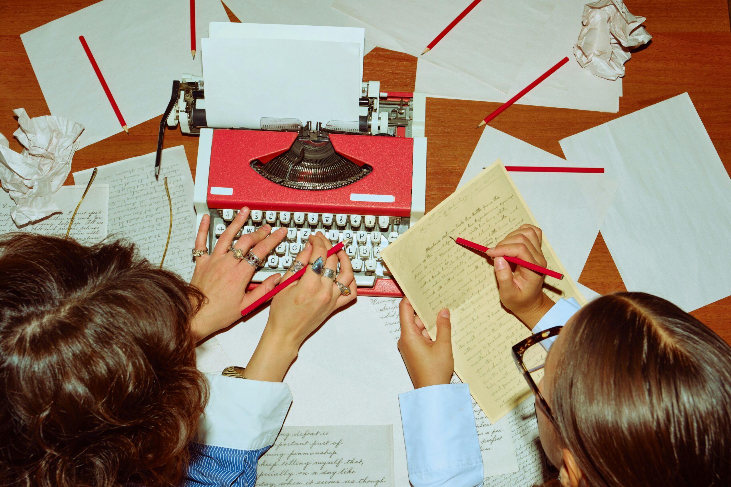 Two people working together at a desk with a red typewriter, handwritten notes, and red pencils.