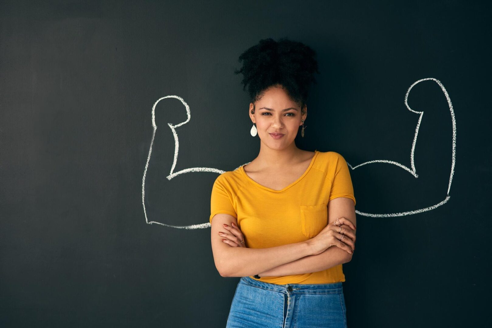 A woman in a yellow shirt stands confidently in front of a blackboard with chalk drawings of flexed biceps behind her, giving the appearance that she has strong arms.