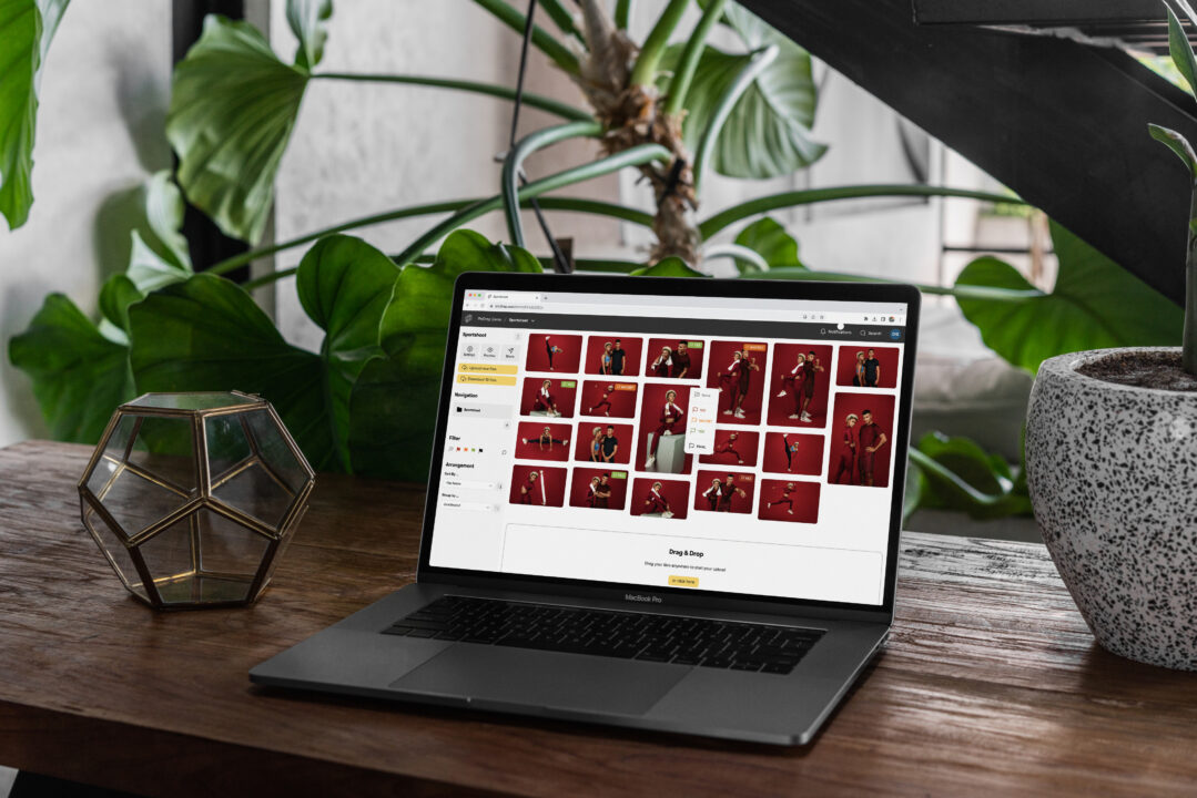 A laptop on a wooden table displays a photo selection screen with thumbnails of portraits in red backgrounds. Plants and a geometric glass decor piece are also visible on the table.