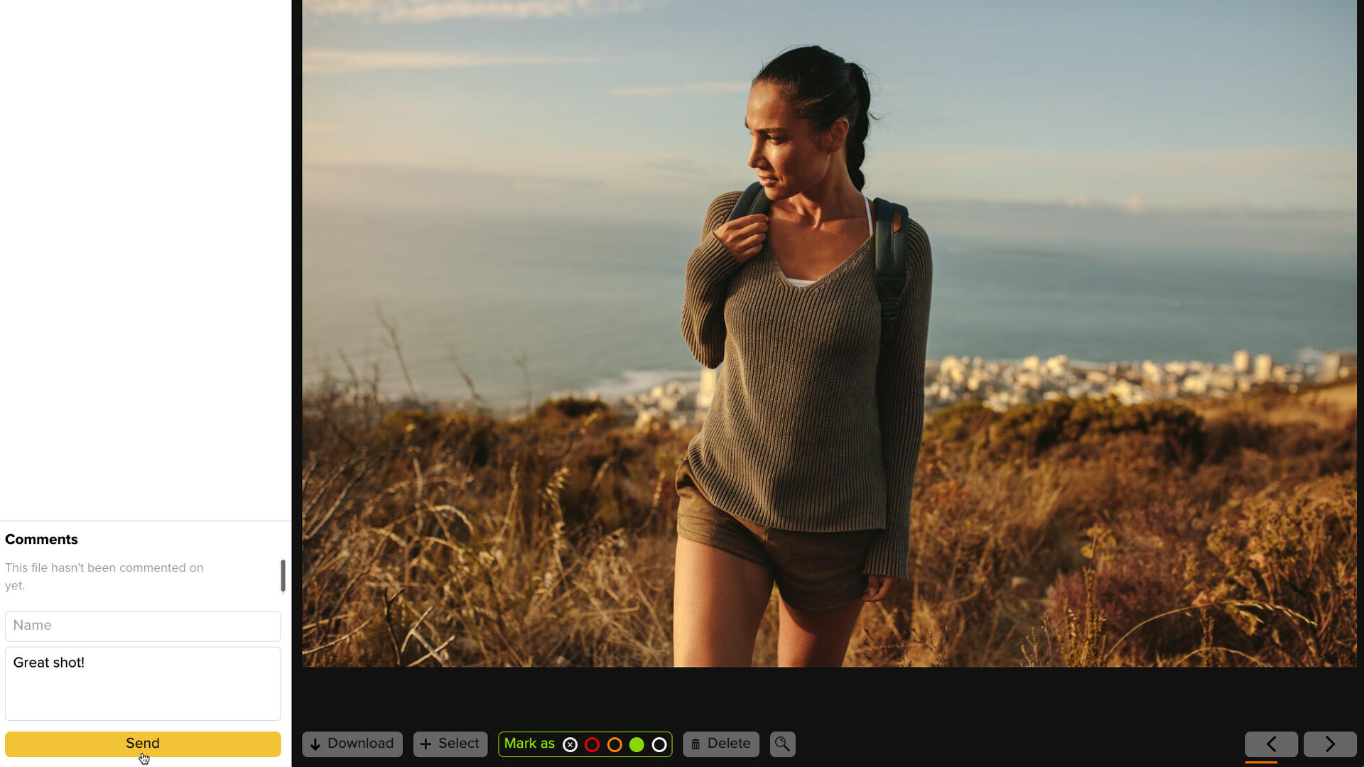 A woman with a backpack walks through dry grass on a hillside overlooking a coastal city and the ocean at sunset. She is wearing a grey sweater and shorts, looking down with a slight smile.