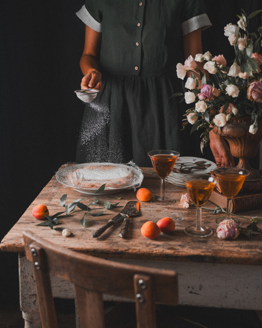 Person sifting powdered sugar over a dessert on a rustic table set with glasses, apricots, flowers, and tableware.