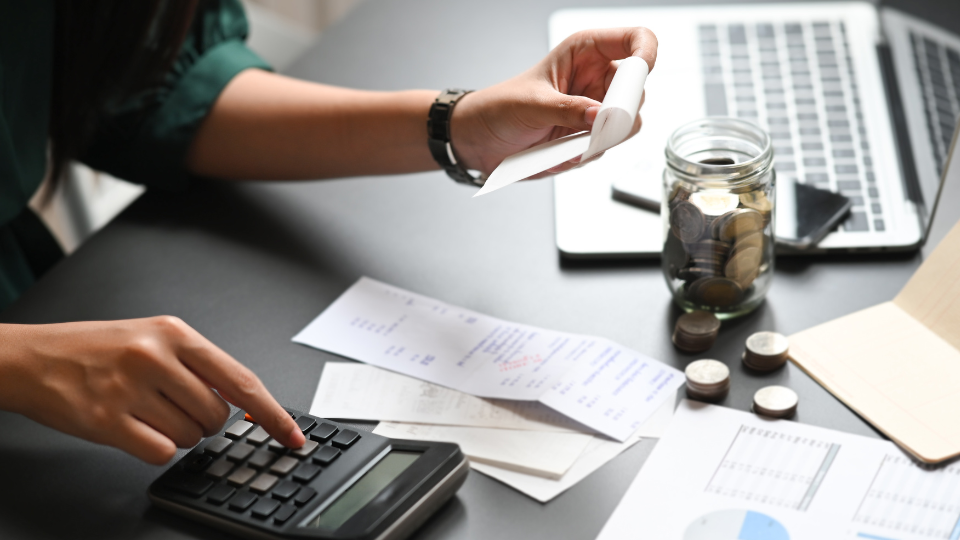 A person uses a calculator while holding a receipt, with papers, a jar of coins, and a laptop on a desk, suggesting budgeting or financial planning activities.