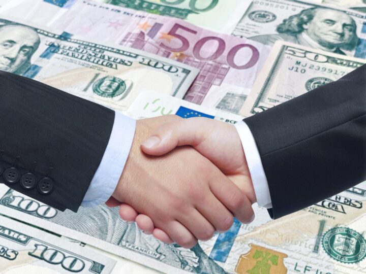 Two people in business suits shaking hands over a background of US dollar and euro banknotes, symbolizing a financial agreement or business partnership.