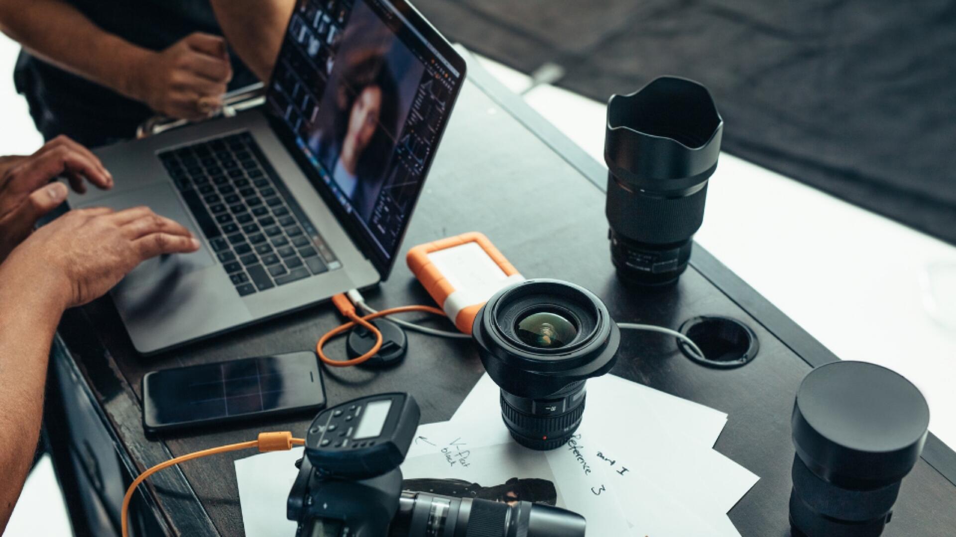 A person works on a laptop at a desk with camera lenses, a DSLR camera, a phone, an external hard drive, and papers scattered around. Another persons hand is visible nearby.