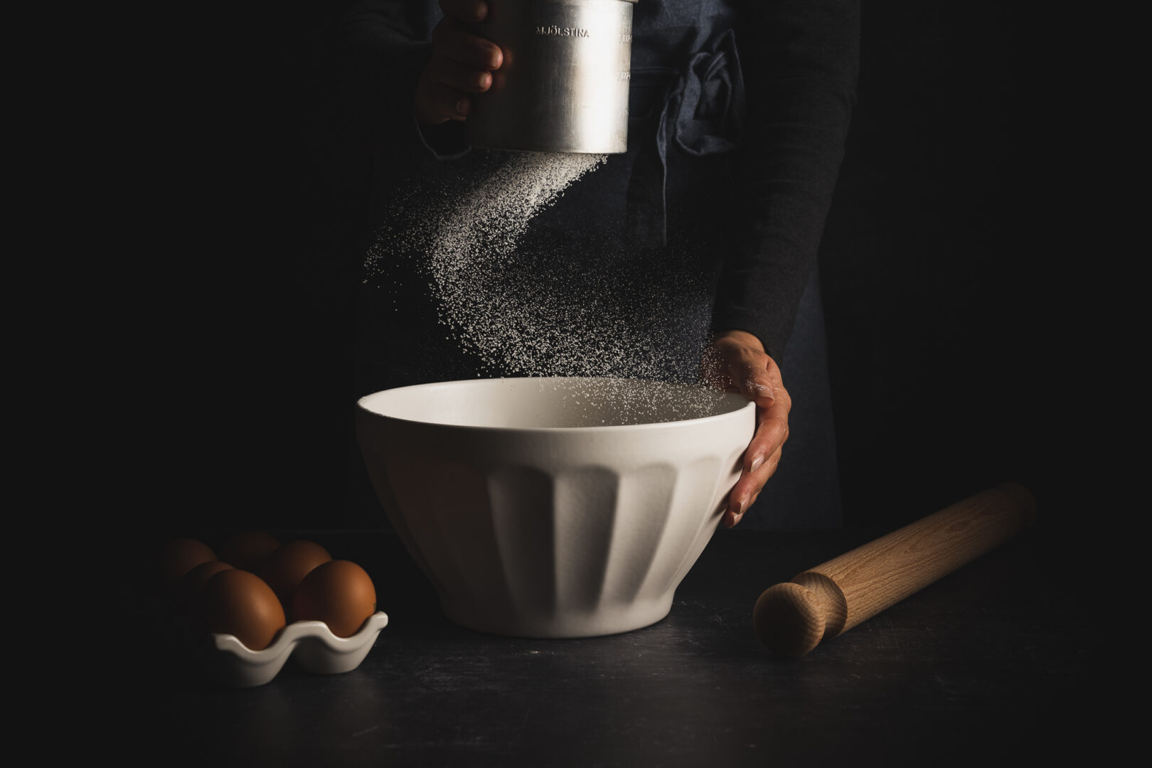 Person sifting flour into a white mixing bowl, with eggs and a rolling pin on a dark work surface.