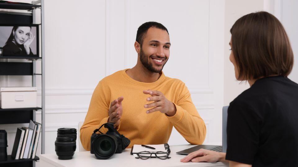 A smiling man in a yellow sweater sits at a desk with a camera, lens, and eyeglasses, talking to a woman across from him in an office with shelves and black-and-white photos in the background.