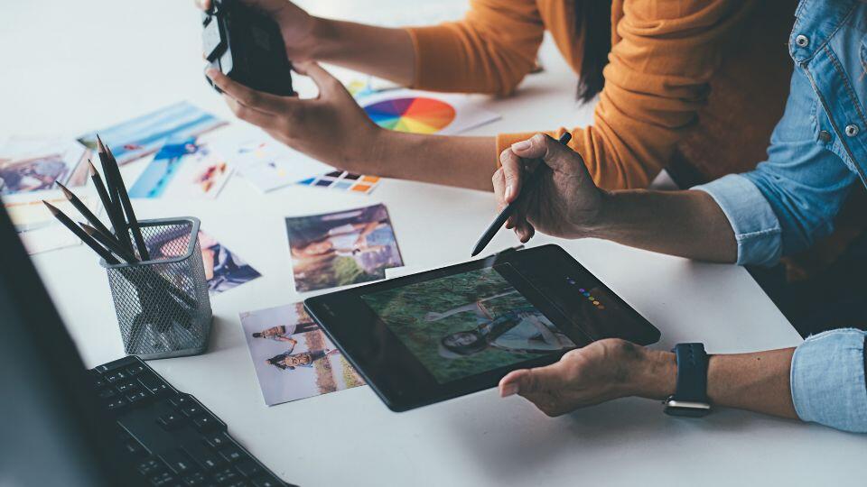 Two people edit a photo on a tablet using a stylus. One holds a camera and the other uses the tablet. Photo prints, color swatches, and pencils are scattered on the white desk. A keyboard is visible in the foreground.