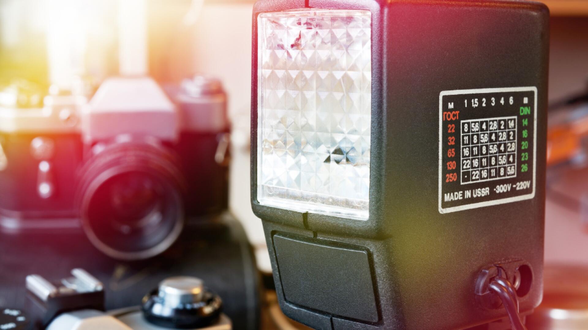 Close-up of a camera flash unit with a settings chart, with blurred vintage film cameras and photography equipment in the background. Bright sunlight is shining on the scene.
