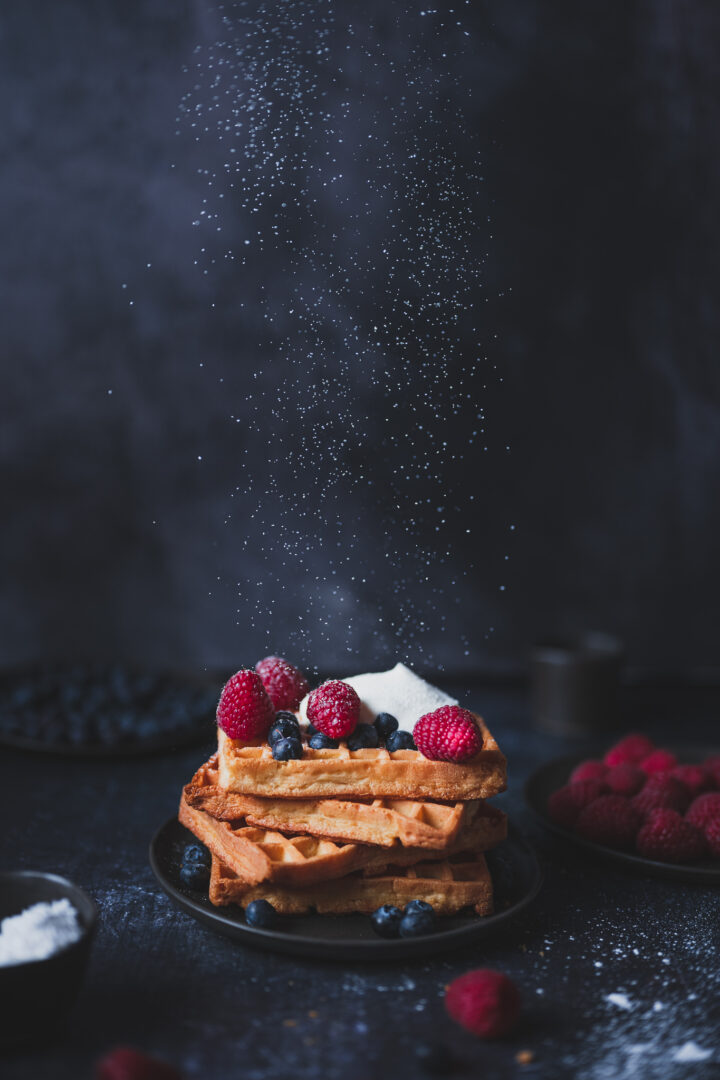 Stack of waffles topped with raspberries, blueberries, and powdered sugar, with more powdered sugar falling through the air against a dark background.