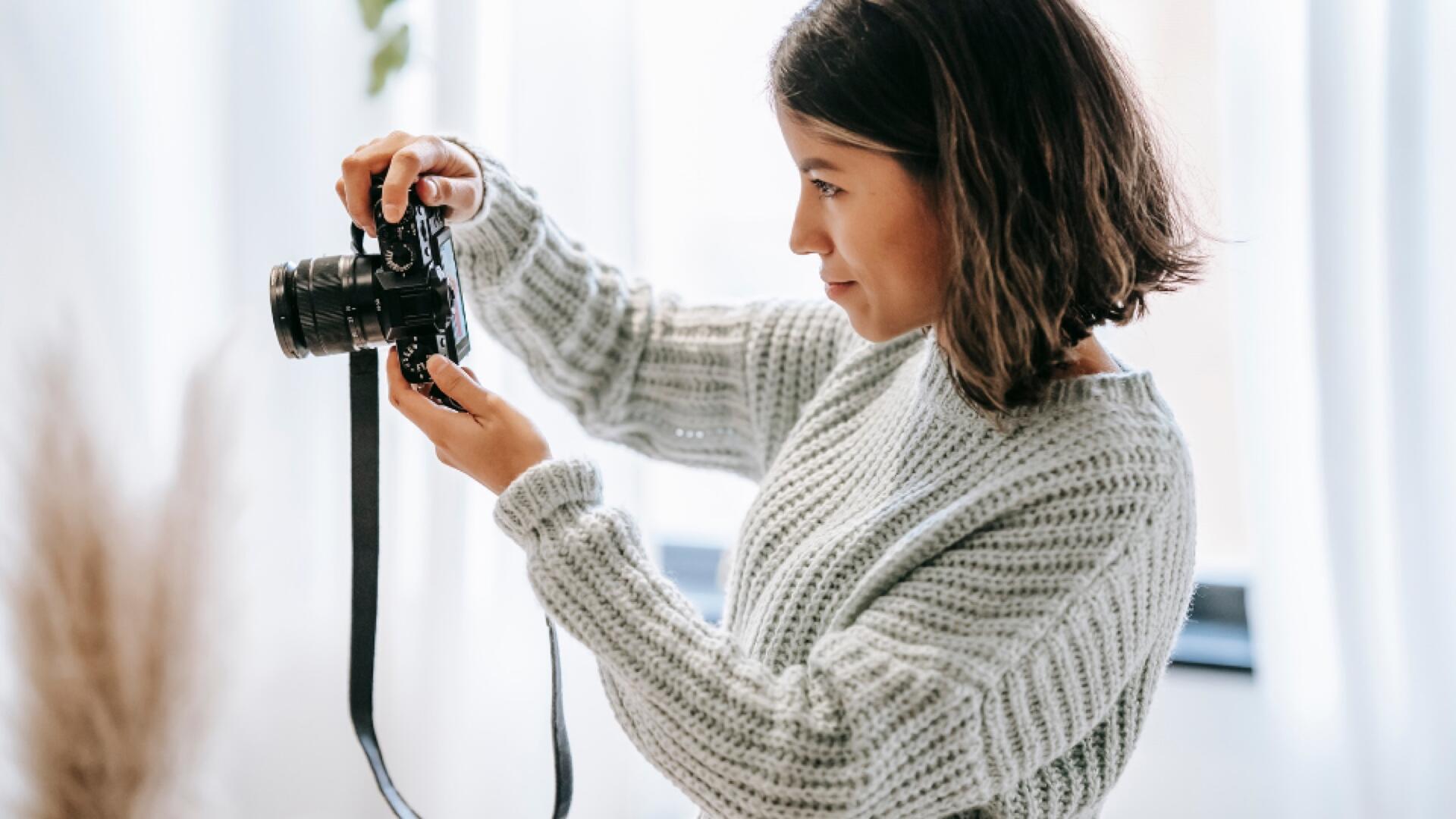 A woman wearing a light gray knitted sweater is holding and adjusting a camera, looking at the controls with focus. She stands indoors in a brightly lit room with a blurred background.