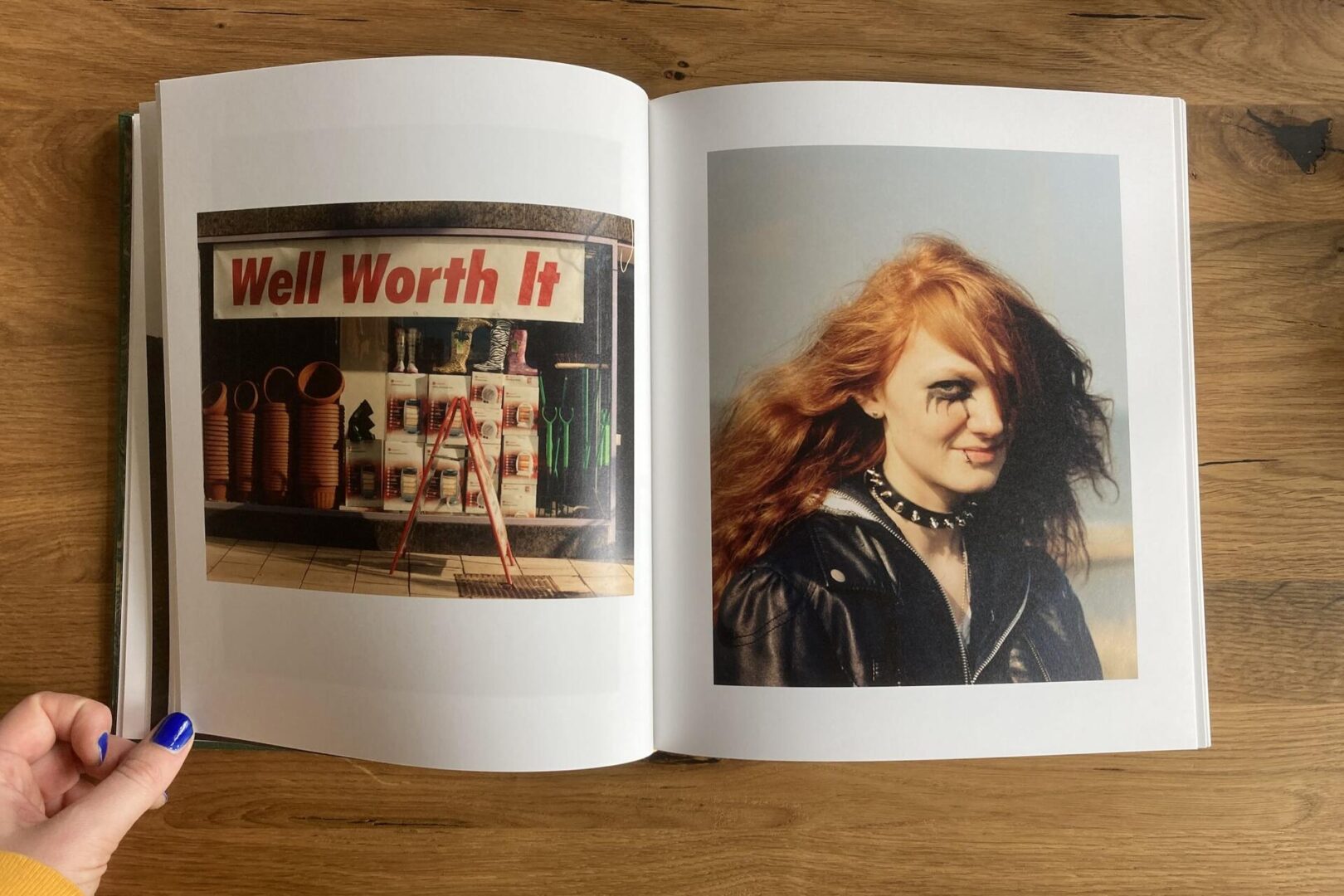 A hand holds open a book showing two pages: the left has a photo of a store display with a Well Worth It sign; the right has a portrait of a person with red hair, heavy makeup, and a spiked choker.