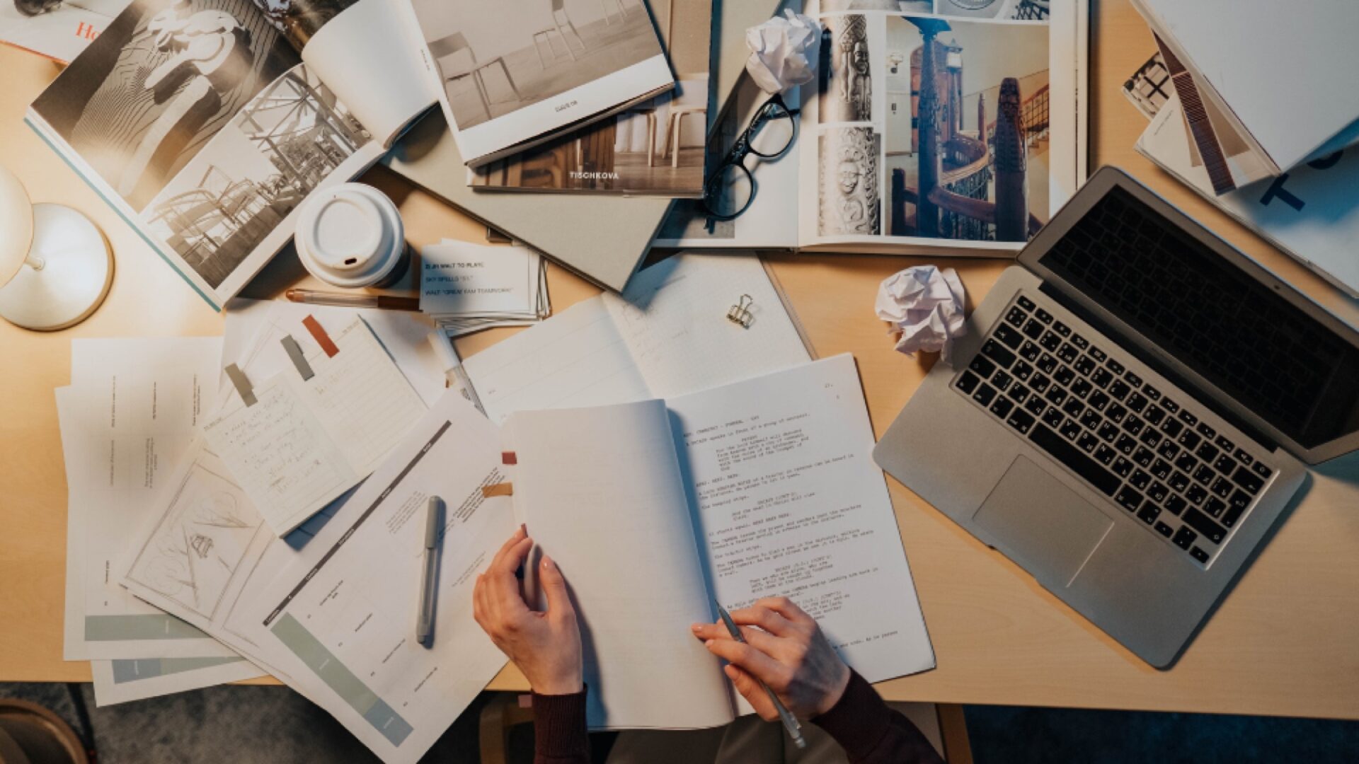 A person studies at a cluttered desk with open books, papers, a laptop, coffee cup, glasses, and crumpled notes, creating a busy and focused workspace atmosphere.